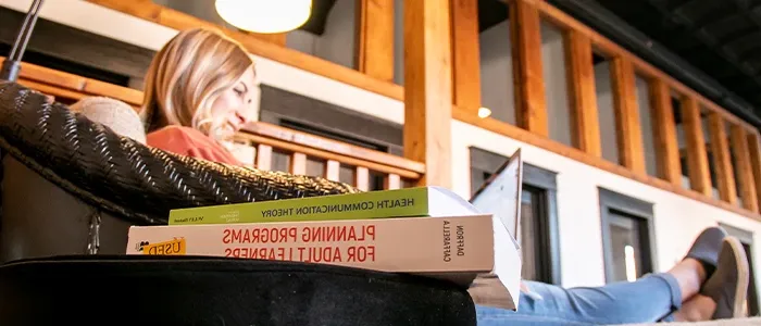 a student lounges while working at her laptop, a pile of communication textbooks sits in the foreground